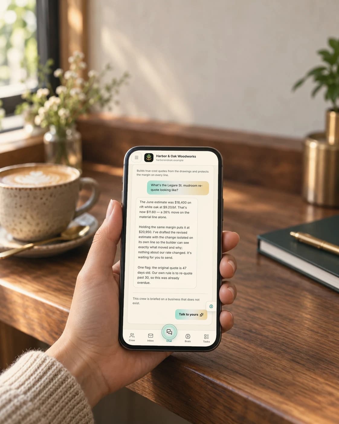 A hand holds a phone showing the Brainztem chat, with a reply about a re-quote waiting to be sent; a latte and a notebook sit on the wooden table beside it.